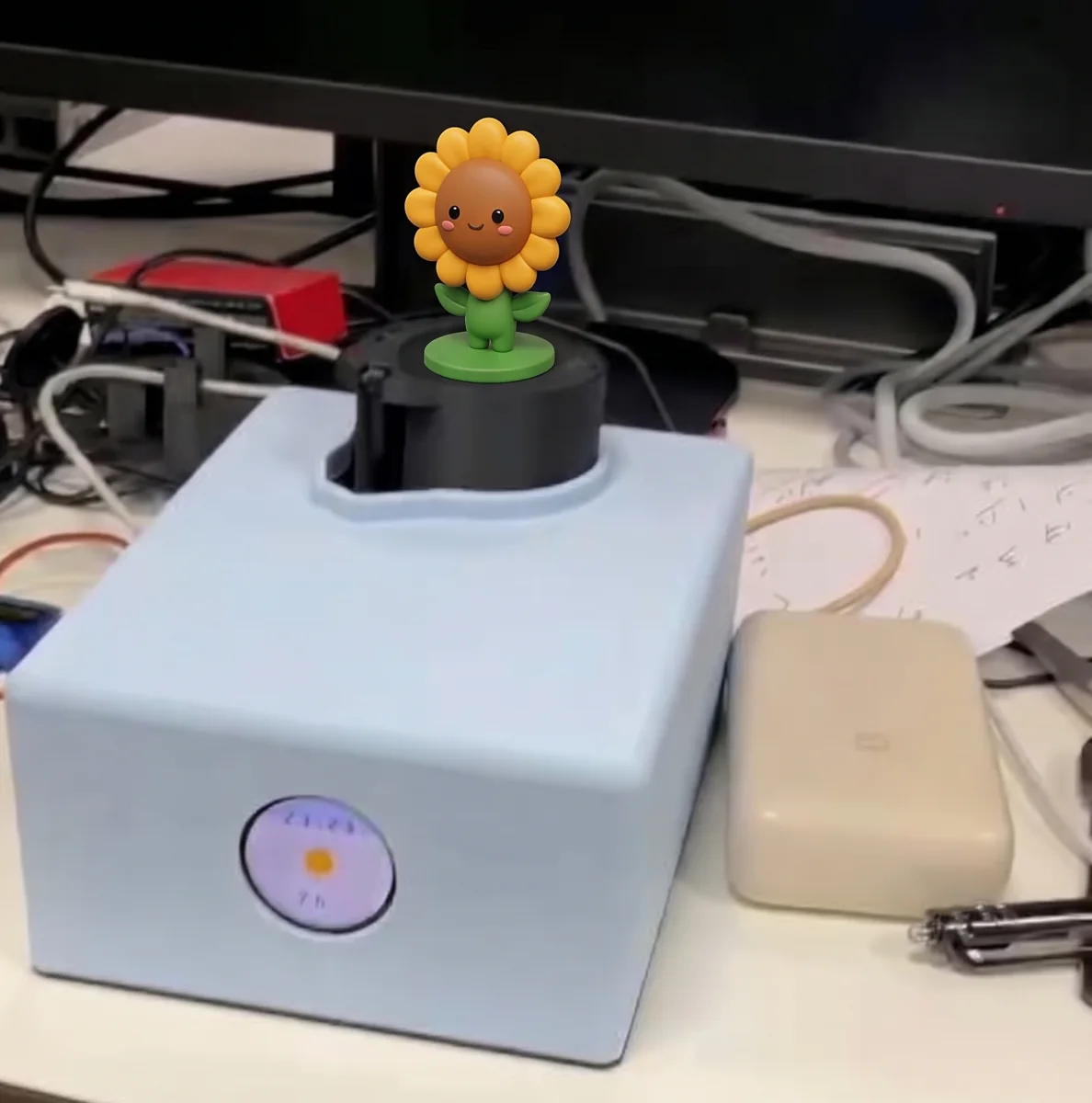 Radar Desk Buddy on a desk showing the clock screen, with a sunflower figure on its servo turntable (illustration)